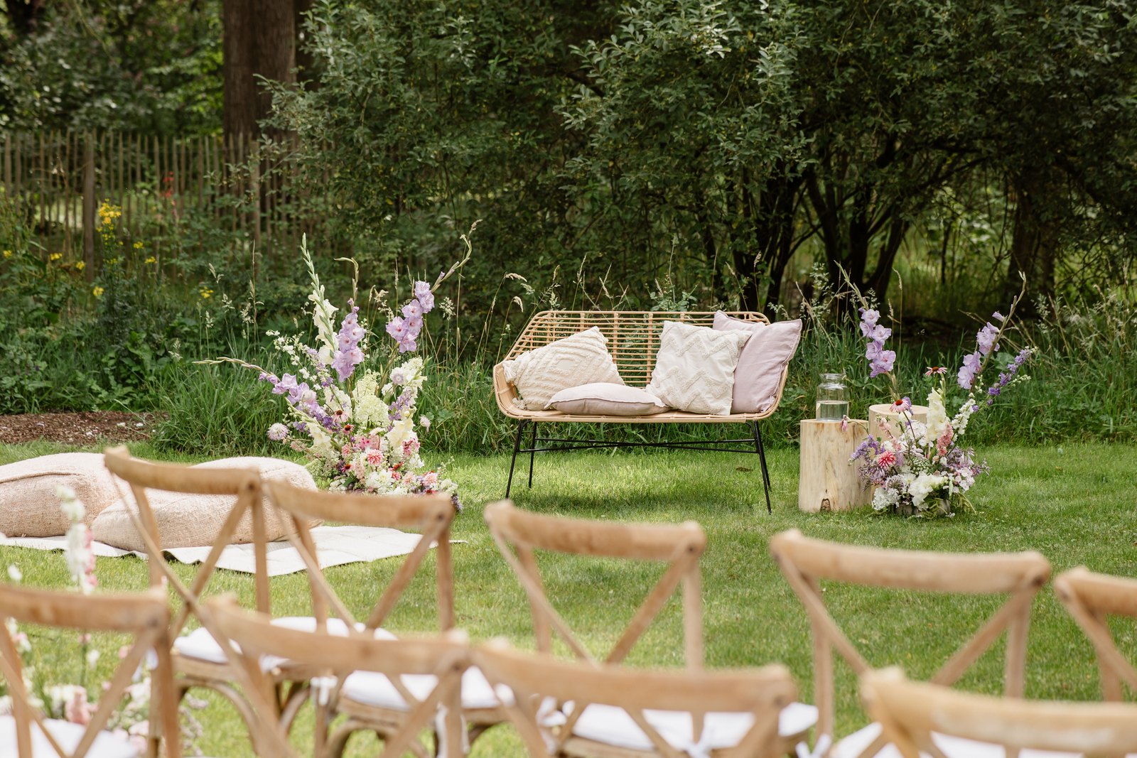 Ceremonieopstelling in de tuin met rieten bank, kussens en lila zomerbloemen