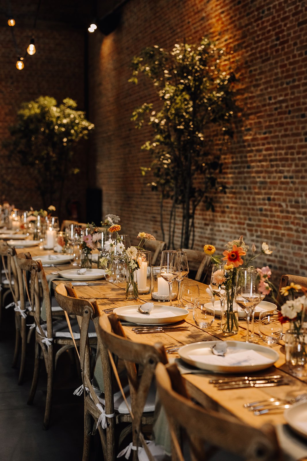 Lange feesttafel met kaarsen en veldbloemen in een schuur met bakstenen muur