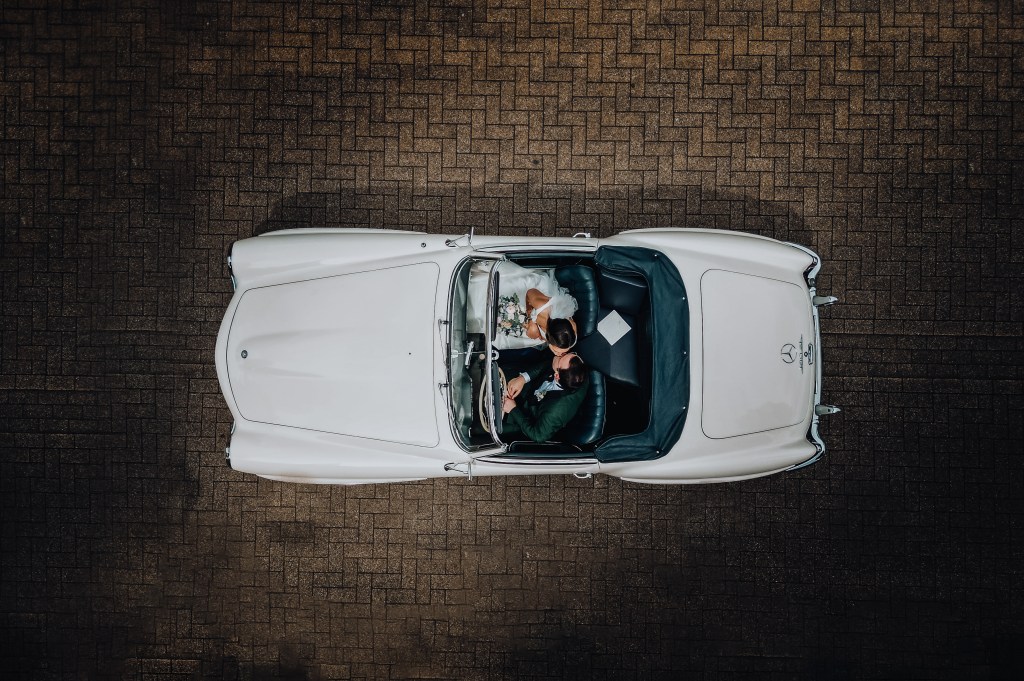 Een koppel kussend in een witte oldtimer. Deze foto is vanuit bovenaanzicht.